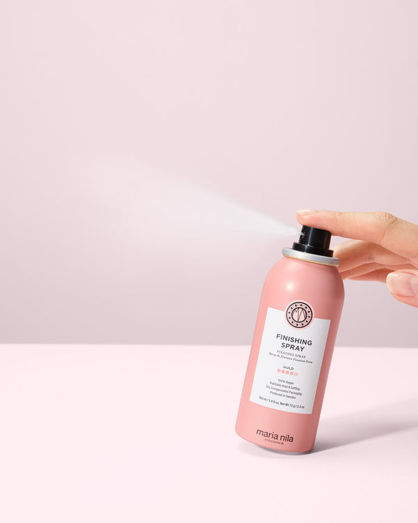 Hand holding a travel size Finishing Spray bottle against a light pink background