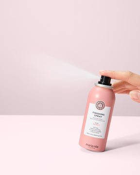 Hand holding a travel size Finishing Spray bottle against a light pink background