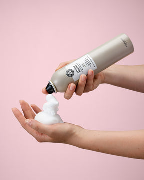 Person holding a bottle of Styling Mousse and pouring it onto their hand against a pink background