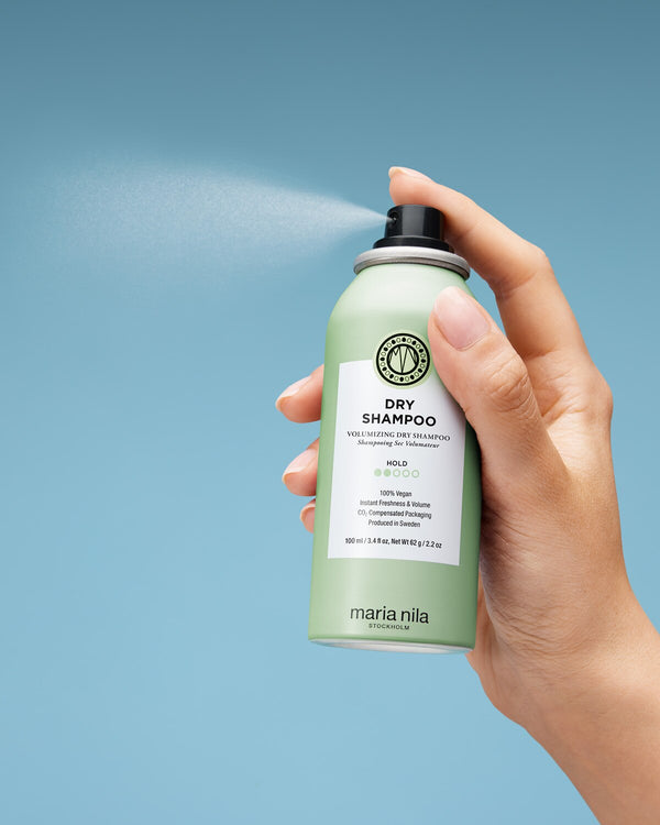 Hand holding a green Maria Nila dry shampoo can against a light blue background