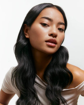 Woman with long, wavy black hair wearing a white off-shoulder top on a light background